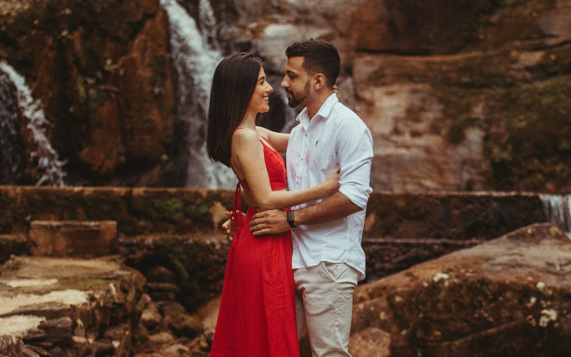 Vista de frente de casal apaixonado abraçados e trocando olhares sob um fundo de pedras e cachoeira.