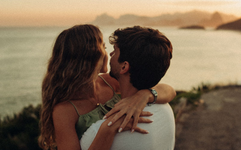 Vista de frente de casal apaixonado se abraçando e olhando para o mar ao fundo da imagem.