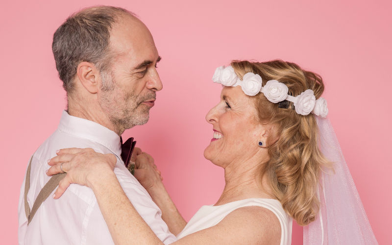 Vista de frente de casal de noivos idosos apaixonados abraçados e se olhando sob um fundo de cor rosa pastel.