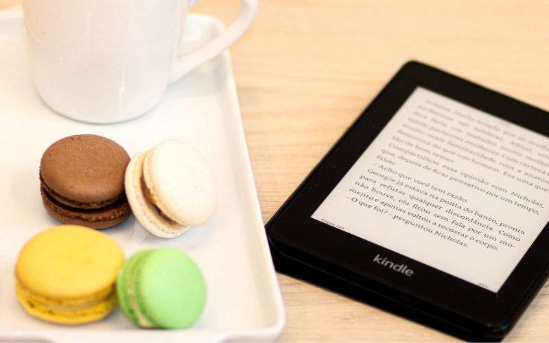 Vista de frente de Kindle posicionado sob uma superfície de madeira ao lado de bandeja com macarons e xícara.