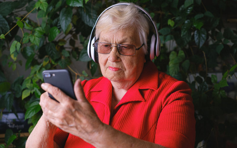 Vista de frente de senhora usando celular com fone de ouvido sem fio em um espaço externo com folhas verdes ao fundo.