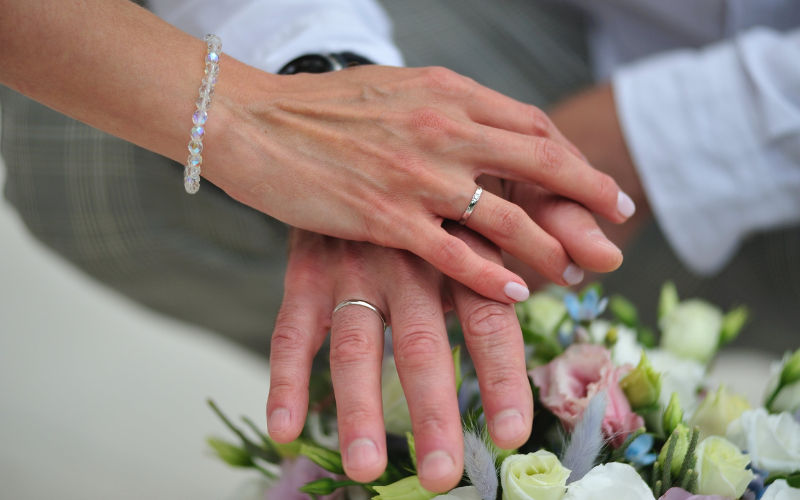 Vista de frente das mãos de um casal com alianças sobrepostas em cima de um buque de flores