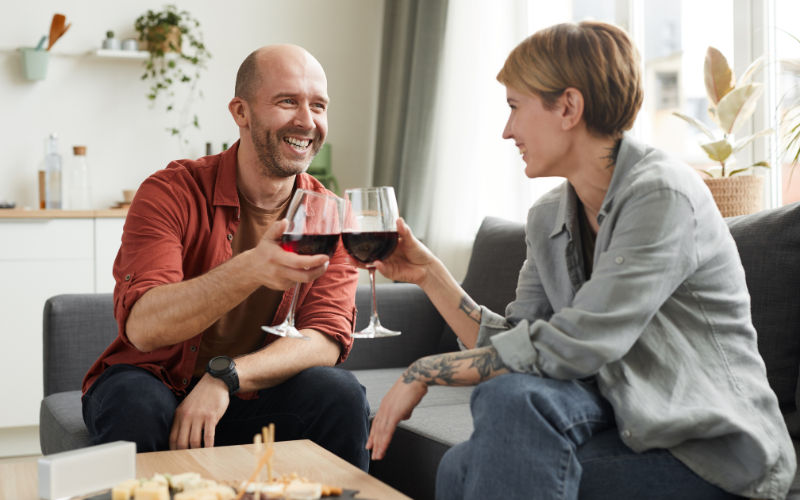 um casal sorridente sentado um sofá brindando com taças de vinho