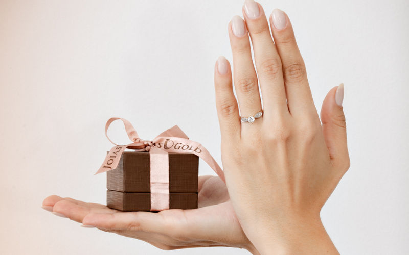 Vista de frente de duas mão femininas em um fundo branco, uma esta segurando uma caixa de joias e a outra exibe um anel